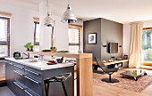 Küche mit Mittelblock und minimalistischer Theke aus Holz, gegenüber Lounge mit Sessel und Bodentisch auf Teppich in modernem, offenem Wohnraum