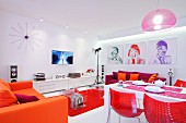Open-plan living-dining room with bright red and orange elements in white interior and green, red and purple portraits on wall