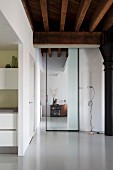 View through floor-to-ceiling, glass sliding door in renovated loft apartment with rustic, wood-beamed ceiling and pale grey resin floor