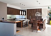Dining set with upholstered rattan chairs, shell stools at counter with sink and open-plan fitted kitchen with exotic-wood fronts and retro lamps above tables