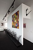Kunstobjekt auf schwarzem Boden, vor Wand mit modernem bunten Bild, an Decke Licht-Schienensystem mit Strahlern