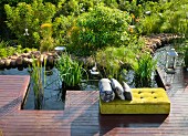 Yellow velvet cushion lounger with grey scatter cushions on wooden deck next to idyllic pool with pebbly shore and reeds
