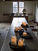 Burning candles on metal trays on a long wooden table with a table under a window in the background in a rustic, historic setting