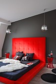 Dramatic bedroom; animal-skin blanket and mattress on platform upholstered in red with tall headboard against dark grey wall