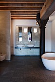Minimalistischer Badbereich, im Hintergrund weiße Aufsatzbecken, seitlich freistehende Badewanne vor schwarzer Metallstütze unter Holzdeckenkonstruktion