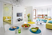 White, open-plan interior with various splashes of colour, dining table, open sliding doors and view into bedroom