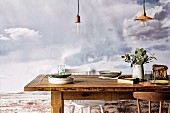 Rustikaler Tisch mit Tellerstapel und Vase, unter Vintage Pendelleuchten, im Hintergrund Bild mit grauer Wolkenstimmung