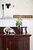 Animal skull and sprig of flowers in apothecary bottle on exotic-wood cabinet against white wainscoting below vintage telephone mounted on ornamental metal wall tiles