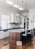 Fitted counter with marble worksurface on Colonial-style wooden base and upholstered bar stools below pendant lamps in elegant country-house kitchen