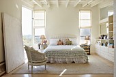Rococo-style chair in front of double bed with ruffled bedspread in bright bedroom with white wooden ceiling