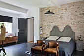 Vintage chairs at foot of double bed with curved, wooden headboard against stone wall