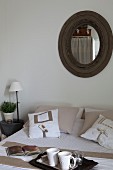 Breakfast tray on bed with arranged scatter cushions below round mirror with rustic wooden frame