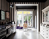 Kitchen with black and white striped wallpaper, modern kitchen counter and retro dresser painted white; dining area with cantilever chairs in background