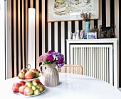 Fruit on cake stand and vase of flowers on white table in front of cylindrical standard lamp against black and white striped wallpaper