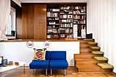 Blue easy chairs in front of raised platform, floor cushions on wooden steps and simple fitted bookcase