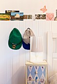 Table lamp on painted bedside cabinet below caps hung on white wainscoting with row of postcards on top