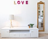Postmodern table lamp on low white sideboard below pink decorative letters spelling 'LOVE' next to gilt-framed mirror
