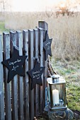 Star-shaped signs made from black-painted wood hanging on a fence with a lantern with a candle on a stone