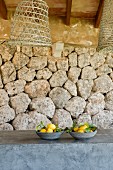 Zwei graue Keramikschalen mit Zitronen auf grauer Mauer, Hängeleuchten mit geflochtenem Lampenschirm vor Wand mit geschichteten Natursteinen
