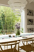 Dining area with pale wooden classic chairs, Enigma designer pendant lamp and view of bamboo bush through window in background