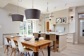 Classic chairs with white shell seats at wooden table below pendant lamps with grey lampshades in front of island counter and bar stools in open-plan, country-house kitchen