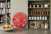 Organised spice jars and tea caddies on olive green kitchen shelves next to red artwork and fruit bowl in open-plan interior