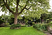 Deciduous tree, flowering herbaceous border, wooden artwork and black outdoor furniture in summery garden