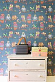 Portable, retro radio and cardboard boxes on vintage chest of drawers against wallpaper with pattern of owls
