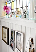 Pictures on white, wood-clad wall below stained-glass transom window with tealight holders and glass dishes on windowsill