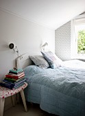 Double bed with white headboard below window and stacked books on stool used as bedside table in bedroom