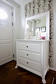 Framed mirror on top of white-painted chest of drawers against leaf-patterned wallpaper
