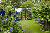 Blue chairs, decorative bottles and blue hydrangeas in garden with greenhouse
