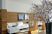 Half-height wood cladding behind white sofa combination, partitions made of bundled brushwood and vase of cherry blossom in foreground