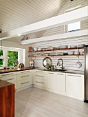 Pale, modern fitted kitchen in renovated country house with crockery on shelves and exposed, white roof beams