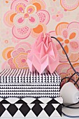 Storage boxes with black and white geometric patterns and table lamp with pink, origami paper lampshade against wall with retro floral wallpaper