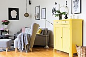 Yellow-painted cabinet next to seating area with grey armchair, matching footstool and retro-style chrome standard lamp