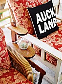 Black and white scatter cushion printed with 'Auckland' motto on floral armchair next to magazine rack