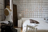 Simple bed with wooden headboard made of branches decorated with fairy lights, vintage typewriter and tailors' dummy in white, wood-clad bedroom