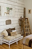 White wicker bench with cushions in various shades of brown next to vintage wooden ladder against white wood-clad wall