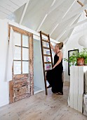 Woman wearing black climbing rustic wooden ladder in country-style summer house with white-painted wood-clad interior