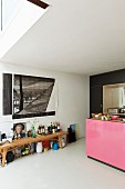 Bottles on wooden bench below drawing on wall and kitchen counter with pink front to one side