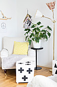 Hell bezogener Hocker mit grafischem Muster vor Sessel, seitlich Zimmerpflanze auf Blumenständer, neben Retro Stehleuchte