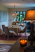 Set dining table and upholstered chairs below chandelier with small lampshades; tealight holders and table lamp providing warm glow in foreground
