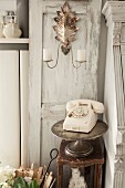 Cream vintage telephone on silver cake stand and old stool below candle sconce on decorative door with patinated paint