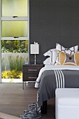 Bedside cabinet next to bed with patterned scatter cushions against wall painted dark grey and next to floor-to-ceiling window with view of garden
