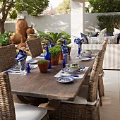 Solid wooden table set in white and blue and wicker chairs on terrace with sofa and terracotta pot in background