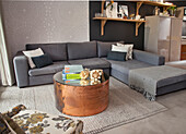 Copper coffee table with mirrored top, grey corner sofa, ornamental wallpaper and ornaments on wooden shelves in open-plan interior