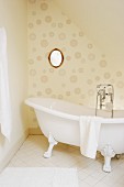 Freistehende Badewanne mit Löwenfüssen und Vintage Armatur vor tapezierter Wand mit kreisförmigem Muster und ovalem Wandspiegel
