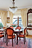 Antique wooden chairs and red and gold upholstery around dining table in grand dining area with pale, draped curtains on French windows