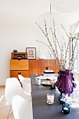 Purple vase of willow branches and tealight holders on table with white classic chairs